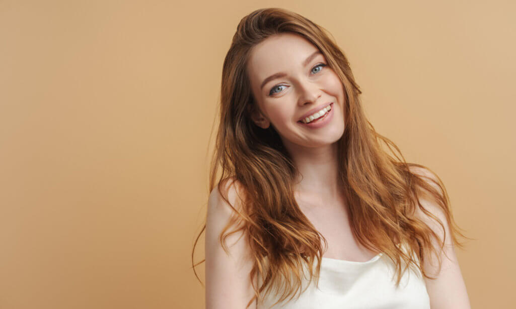 Portrait of smiling positive woman with toothy smile looking at camera isolated on beige background. Beautiful girl with healthy hair posing for picture. Healthy lifestyle concept