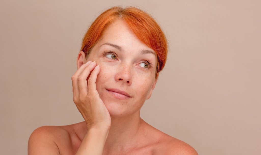 Portrait of cropped caucasian middle aged woman face with freckles and reddish hair holding hands on chin over grey background looking up