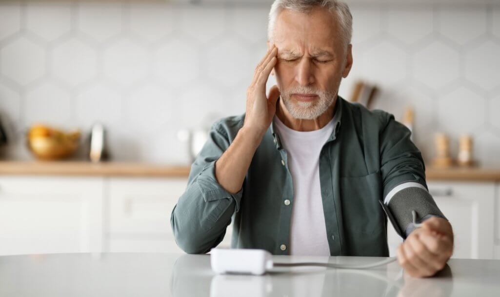 Hypertension Concept. Senior Man Checking Blood Pressure With Upper Arm Monitor At Home, Elderly Gentleman Touching Temples And Measuring Arterial Tension While Sittin At Table In Kitchen