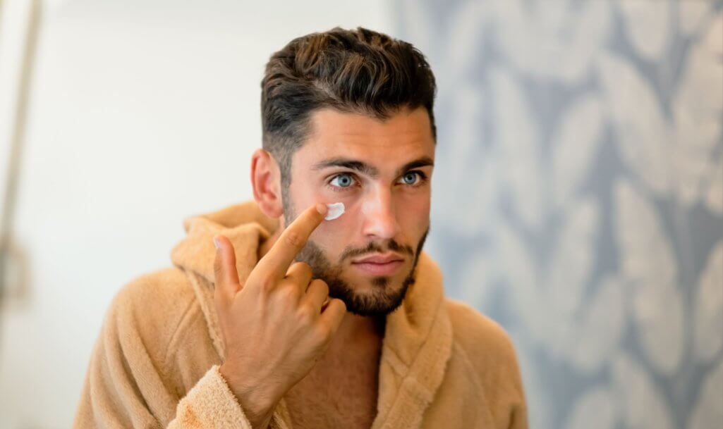 Young bearded male in bathrobe looking at mirror and smearing moisturizing cream under blue eye during skin care routine in morning